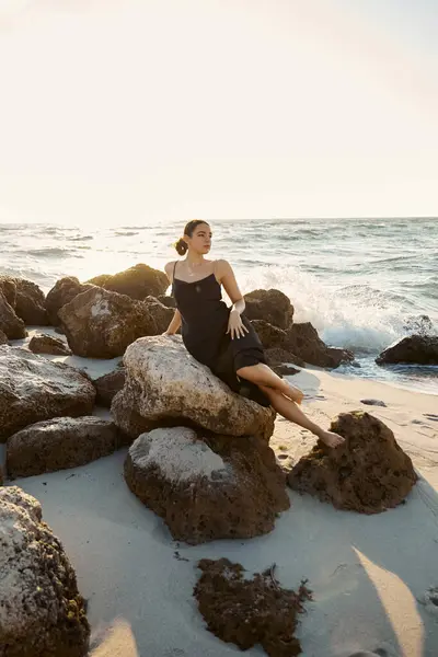 stock image A young woman in a black sundress sits on a rock by the ocean, enjoying the warm Miami sunshine.