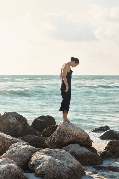 A woman wearing a black sundress stands on a rock formation by the ocean in Miami Beach, Florida.