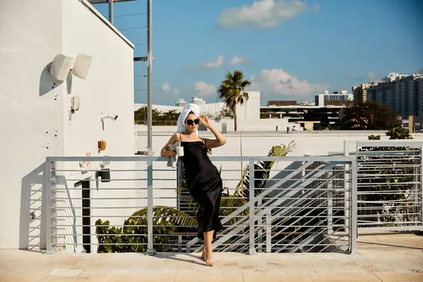 Siyah slip elbiseli ve güneş gözlüklü bir kadın Miami havuzuna bakan bir villanın çatısındaki korkuluklara yaslanıyor..