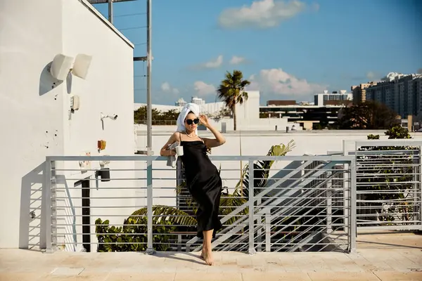 stock image A woman in a black slip dress and sunglasses leans against a railing on a villa rooftop overlooking a Miami pool.