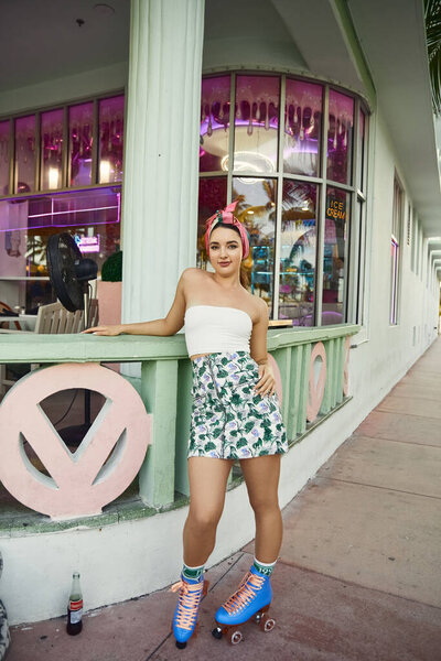 Молодая женщина в солнечных очках и цветочная юбка проезжает мимо винтажного магазина в Майами.