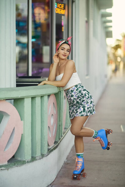 A stylish young woman in sunglasses leans against a railing, one roller skate raised, in Miami.