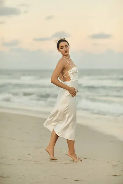 stock image A young woman in a white dress walks along a sandy beach in Miami, with the setting sun casting a warm glow over the ocean.