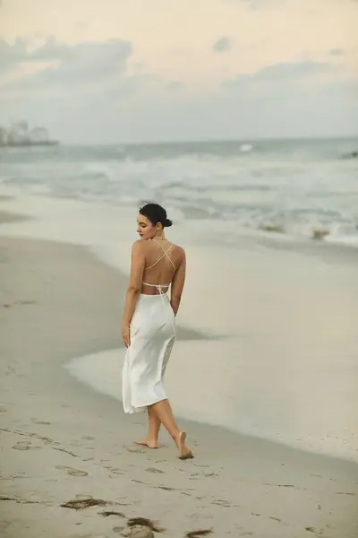 stock image A young woman in a white dress walks along a sandy beach in Miami, her hair blowing in the breeze.