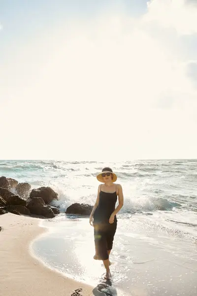 Stock image A woman in a dress walks along a sandy beach in Miami, with the ocean waves crashing behind her.