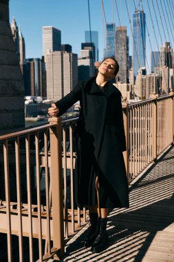 Siyah ceketli genç bir kadın New York şehrinin ufuk çizgisine bakan bir köprüde dans ediyor..