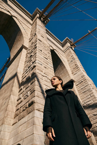 A young woman in a black coat stands beneath the imposing arch of a New York City bridge.