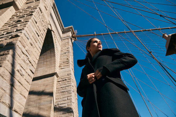 Женщина в черном пальто смотрит на небо у Бруклинского моста.