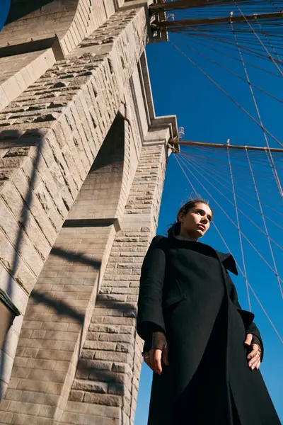 stock image A woman stands beneath the Brooklyn Bridge, her silhouette stark against the clear blue sky.