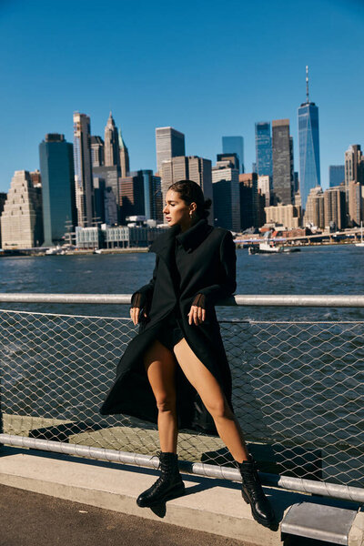 A young woman dances on a New York City pier, wearing a black coat and boots.