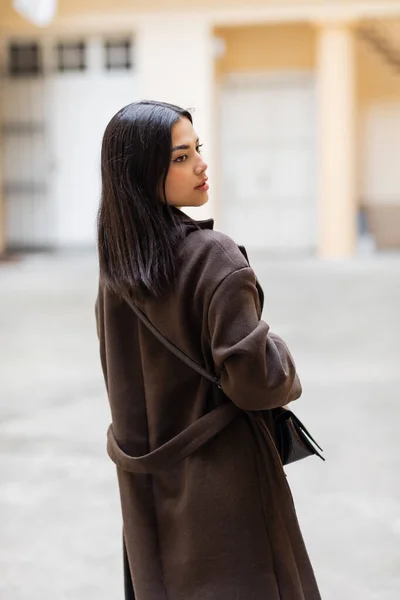 Back view of young brunette woman in trendy coat with crossbody looking away outdoors in prague — Stock Photo