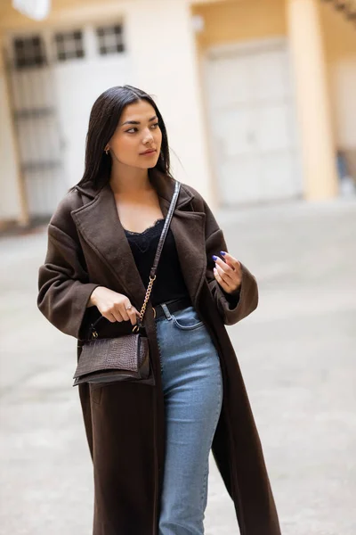 Young woman in coat and jeans holding crossbody on city street in prague — Stock Photo