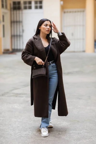 Bella donna bruna in cappotto marrone toccare i capelli e guardando lontano sulla strada urbana a Praga — Foto stock