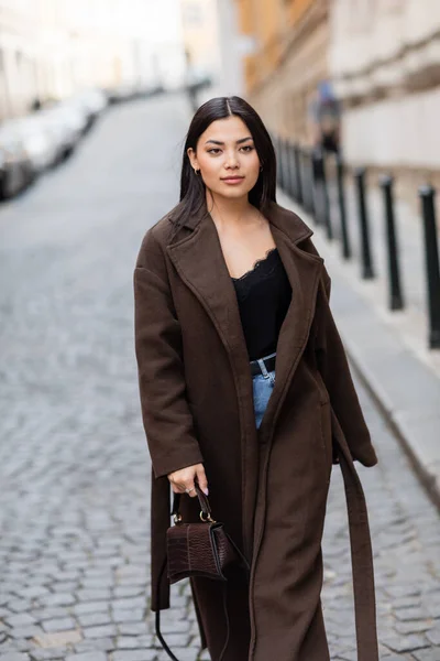 Stylish brunette woman in brown coat holding handbag while walking on street in prague — Stock Photo