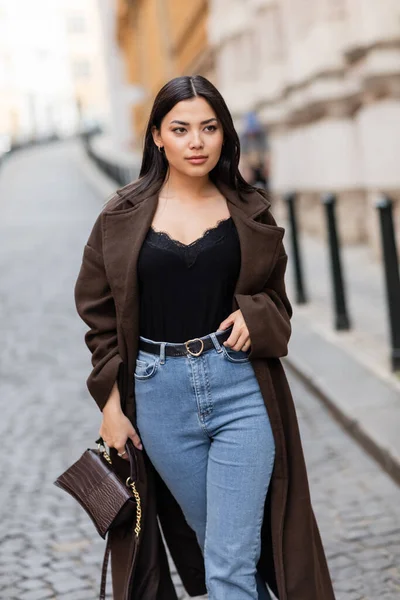 Trendy brunette woman with handbag touching belt of jeans on blurred street in prague — Stock Photo