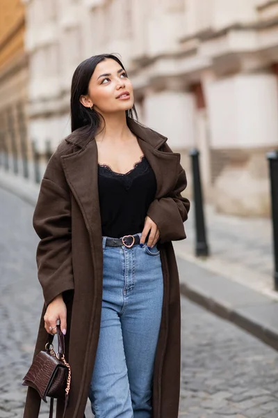Jovem e bonita mulher na moda casaco segurando bolsa e olhando para longe em praga na rua urbana — Fotografia de Stock
