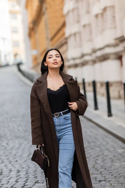 Trendige Frau in braunem Mantel, die mit Handtasche auf der Prager Straße spaziert und wegschaut — Stockfoto