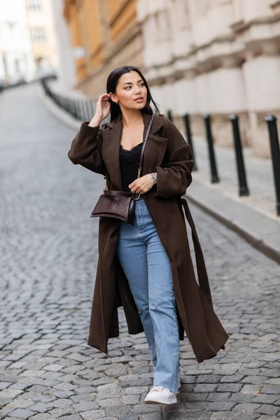 Brunette woman in trendy coat with crossbody fixing hair and looking away on blurred street in prague — Stock Photo