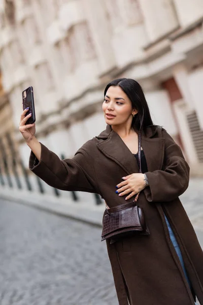 Joven morena en abrigo marrón tomando selfie en calle borrosa en praga - foto de stock