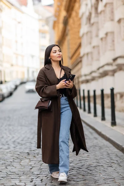 Woman in trendy coat holding smartphone while walking on blurred street in prague — Stock Photo