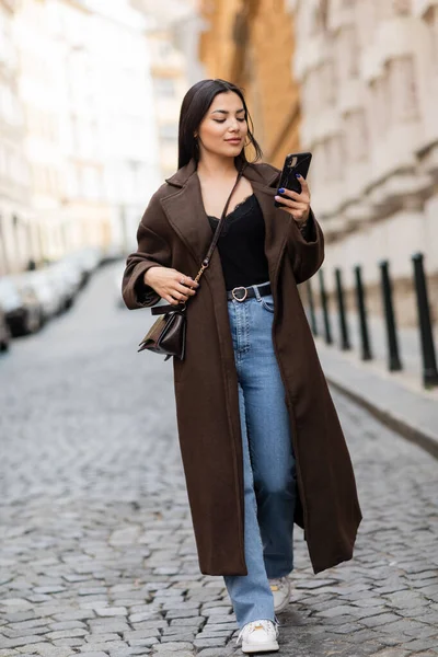 Brunette woman in coat and jeans looking at smartphone while walking on street in prague — Stock Photo