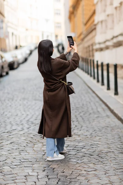 Back view of brunette woman in brown coat taking photo on street in prague — Stock Photo