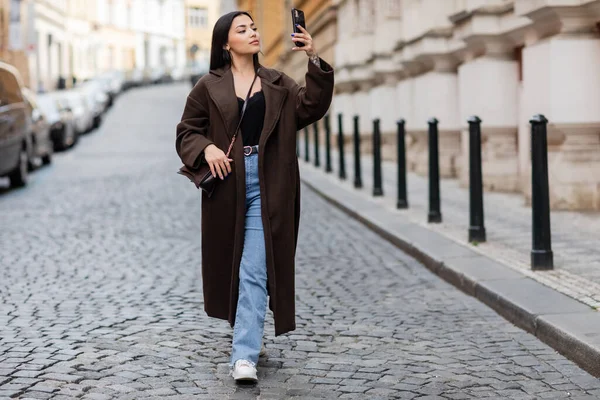 Full length of woman in stylish coat taking photo while walking on pavement in prague — Stock Photo