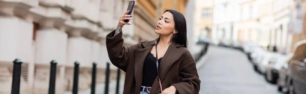 Pretty brunette woman taking photo on cellphone on blurred street in prague, banner — Stock Photo