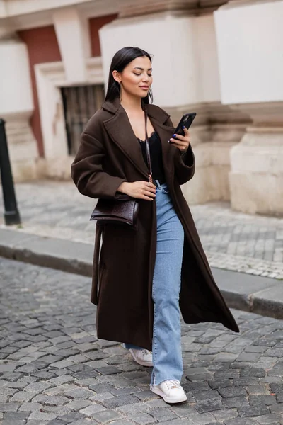 Full length of woman in coat and jeans walking in prague and looking at smartphone — Stock Photo