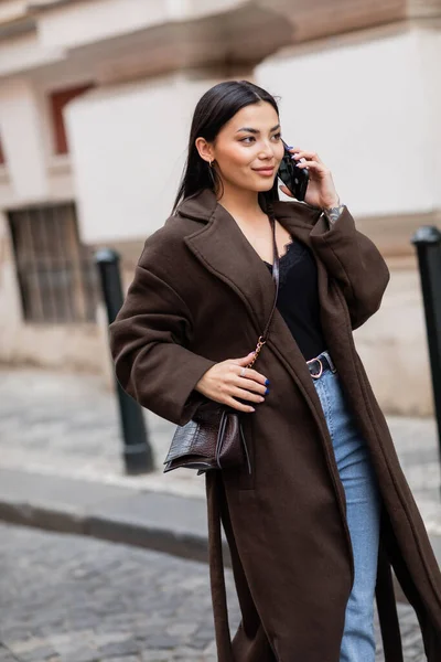 Sonriente morena mujer en elegante abrigo hablando en el teléfono inteligente en la calle borrosa en la praga - foto de stock