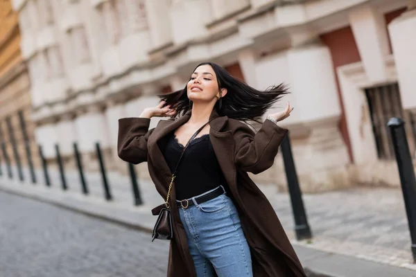 Happy brunette woman in coat and jeans touching hair on urban street in prague — Stock Photo