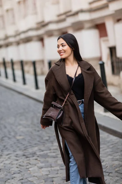 Fröhliche brünette Frau in stylischem Mantel mit Crossbody, die in Prag auf der städtischen Straße spaziert — Stockfoto