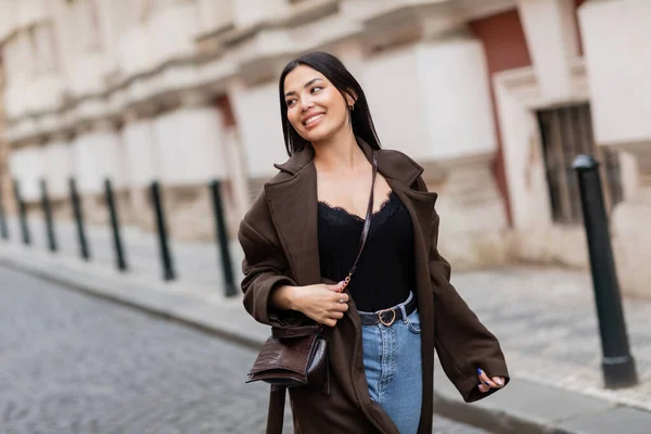 Donna bruna sorridente in cappotto alla moda con crossbody a piedi sulla strada della città a Praga — Foto stock