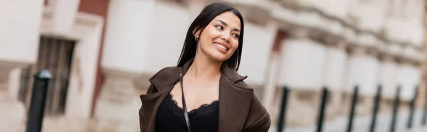 Веселая брюнетка в пальто улыбается на городской улице в Праге, баннер — стоковое фото
