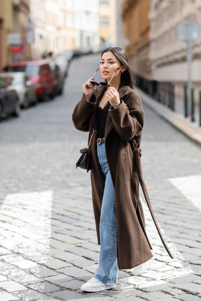 Full length of pretty brunette woman in coat holding sunglasses on city street in prague — Stock Photo