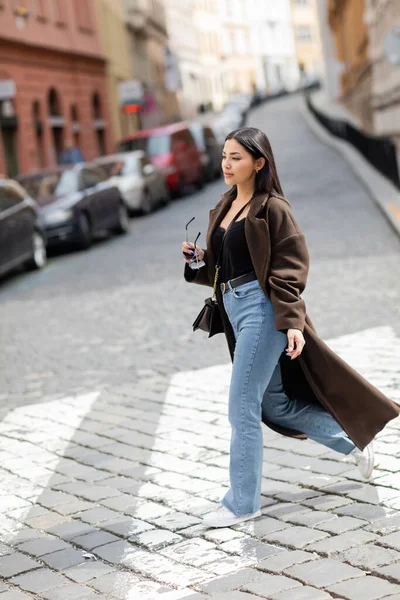 Full length of trendy woman in coat and jeans holding sunglasses while crossing street in prague — Stock Photo