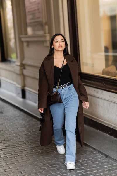 Stylish woman in jeans and coat holding sunglasses while walking along building in prague — Stock Photo