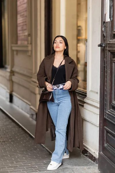 Brunette woman in trendy coat holding sunglasses while standing near building in prague — Stock Photo