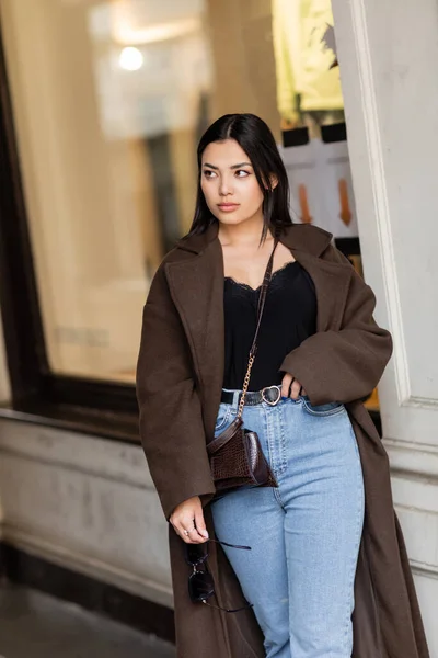 Красивая женщина в модном пальто и джинсах глядя в сторону возле размытого здания на улице в Праге — стоковое фото