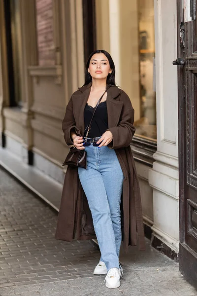Full length of young woman in stylish coat and jeans holding sunglasses near building in prague — Stock Photo