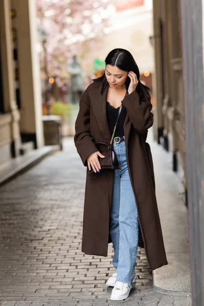 Volle Länge der brünetten Frau im trendigen Mantel mit Crossbody Fixierung Haare auf der Straße in Prag — Stockfoto