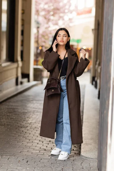 Pleine longueur de femme brune en jeans et manteau parler sur smartphone dans la rue dans la prague — Photo de stock