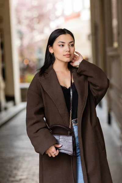 Young woman in brown coat holding hand near face and looking away on street in prague — Stock Photo