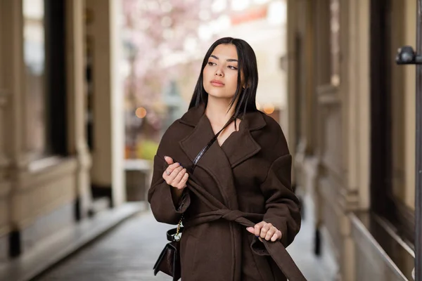 Mulher morena com crossbody amarrando cinto de casaco na moda na rua em prague — Fotografia de Stock