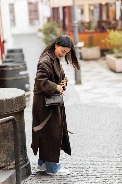 Volle Länge der lächelnden Frau in braunem Mantel, die den Kreuzkörper öffnet, während sie auf der Straße in Prag steht — Stockfoto