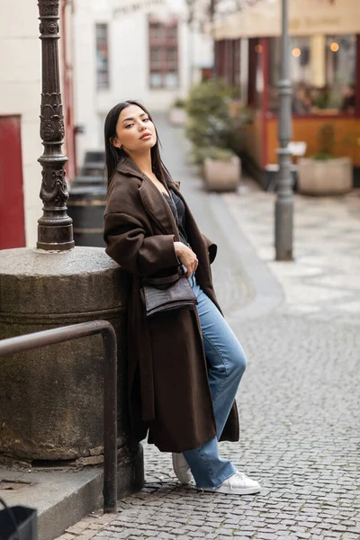 Full length of brunette woman in trendy coat with crossbody looking away near lamppost in prague — Stock Photo