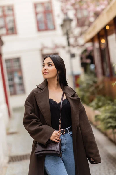 Jolie femme brune en manteau avec bandoulière marchant sur la rue étroite dans la prague — Photo de stock