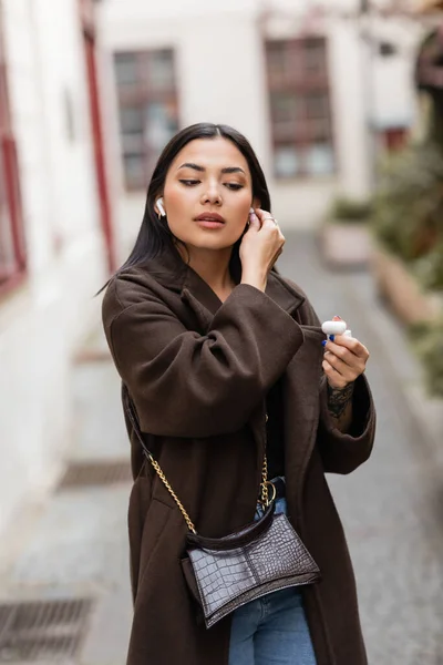 Young woman in trendy coat inserting wireless earphone on blurred street in prague — Stock Photo