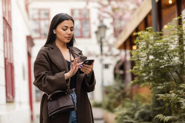 Brunette woman in coat and wireless earphone using smartphone near green plants on street in prague — Stock Photo