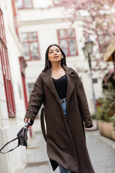 Hübsche brünette Frau in trendigem Mantel und drahtlosen Kopfhörern, die mit Handtasche in Prag spazieren geht — Stockfoto